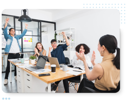 A diverse group of five young adults celebrates in a modern office. They cheer with raised arms around a table with laptops, conveying excitement and teamwork.