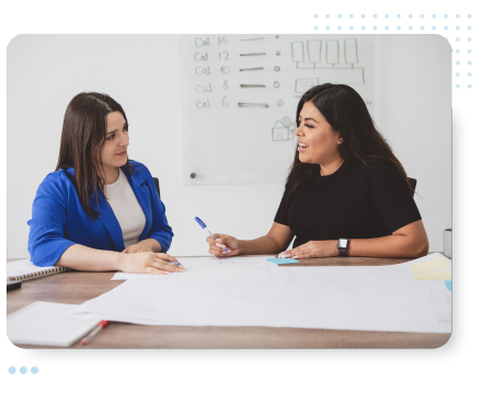 Two women sit at a table, engaged in discussion. One is in a blue jacket, the other in black, both smiling. Papers and a whiteboard are in the background, collaborating to optimize HR services and meet employee needs.